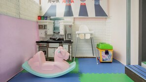 Una sala de juegos con un caballito de madera rosa, una cocina de juguete y un contenedor verde para guardar cosas.