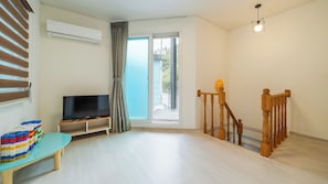 A room with a flat-screen TV on a stand, a window with curtains, a staircase with wooden railings, and a light fixture.