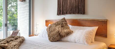 A bedroom with a large bed, a wooden headboard, a macrame wall hanging, and a view of greenery outside.