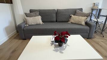 Un sofá gris con cojines estampados, una mesa de centro blanca con flores rojas y una silla de metal.