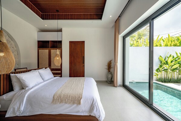 A modern bedroom with a large bed, a wooden ceiling, a glass door leading to a pool, and a view of greenery outside.