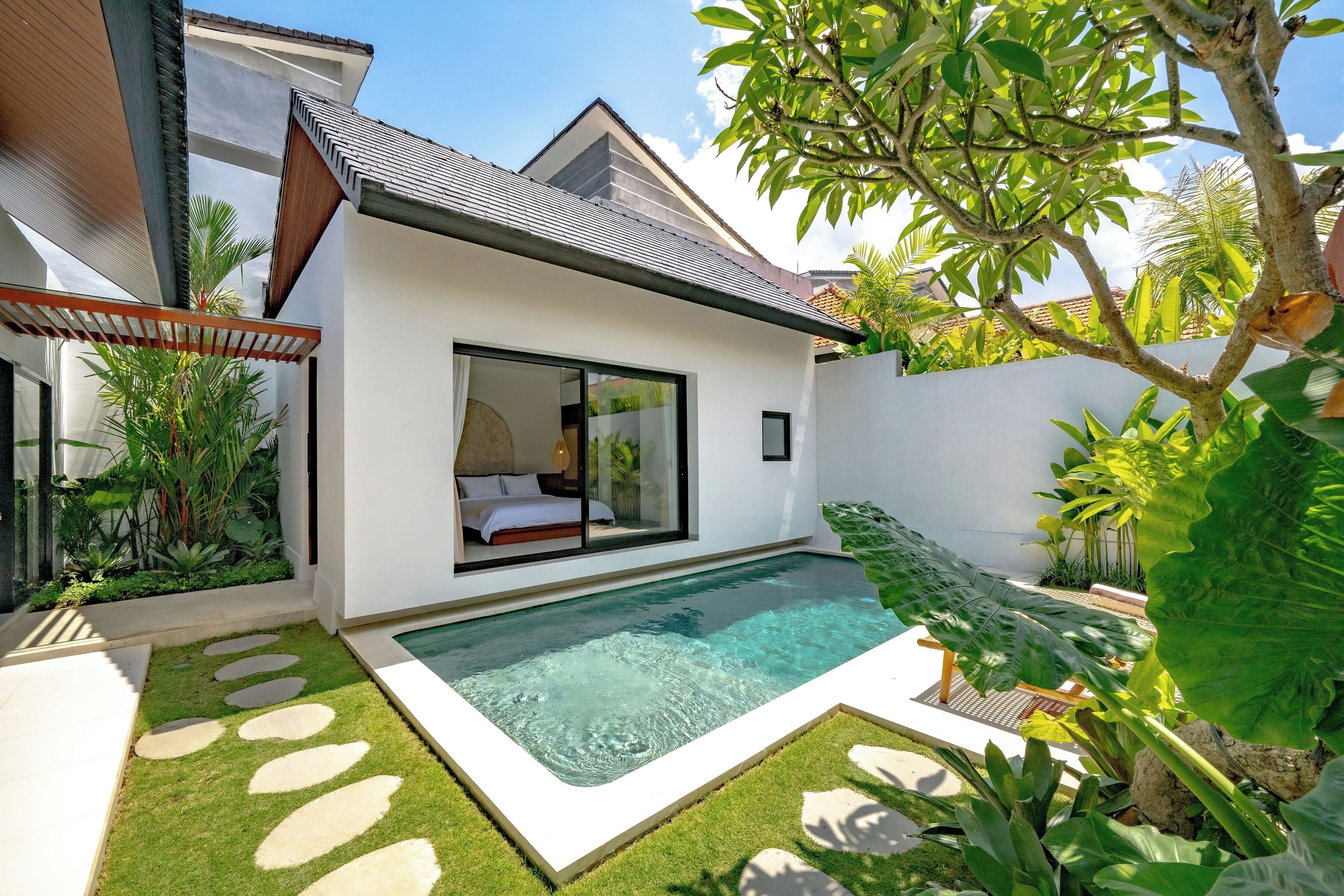 Una casa moderna con piscina, un dormitorio con paredes de cristal y un jardín con plantas tropicales.