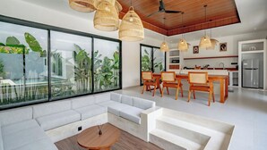 A modern living room with a large window, a sofa, a dining area with wooden chairs, and a kitchen in the background. - Casa Umari (Canggu)