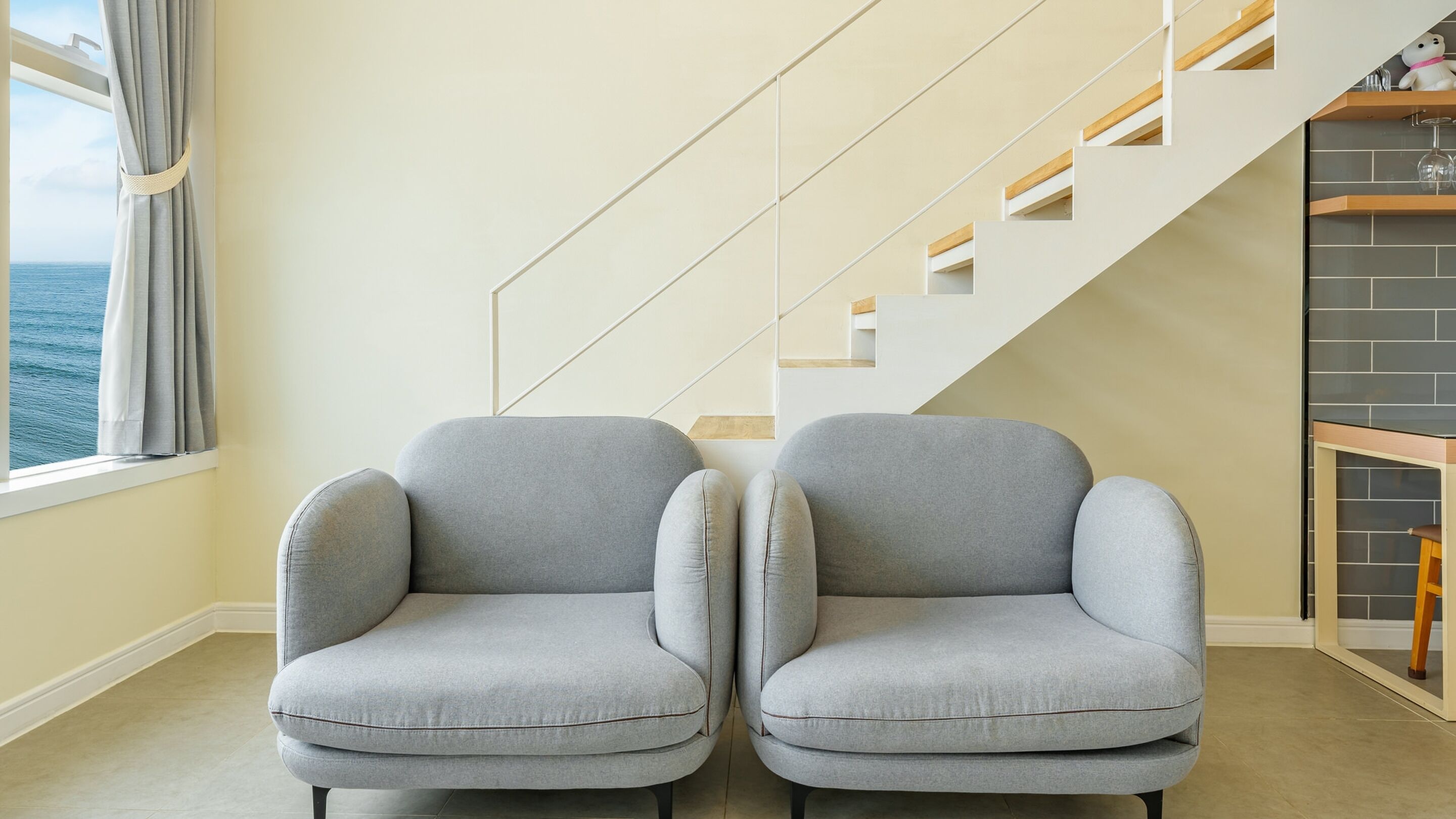 A room with two grey armchairs, a staircase with wooden steps and white railings, and a view of the sea through a window with grey curtains.