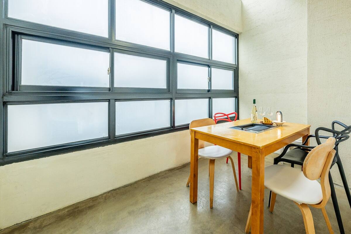 Un comedor con mesa y sillas de madera, una ventana con marcos negros y suelo de hormigón.