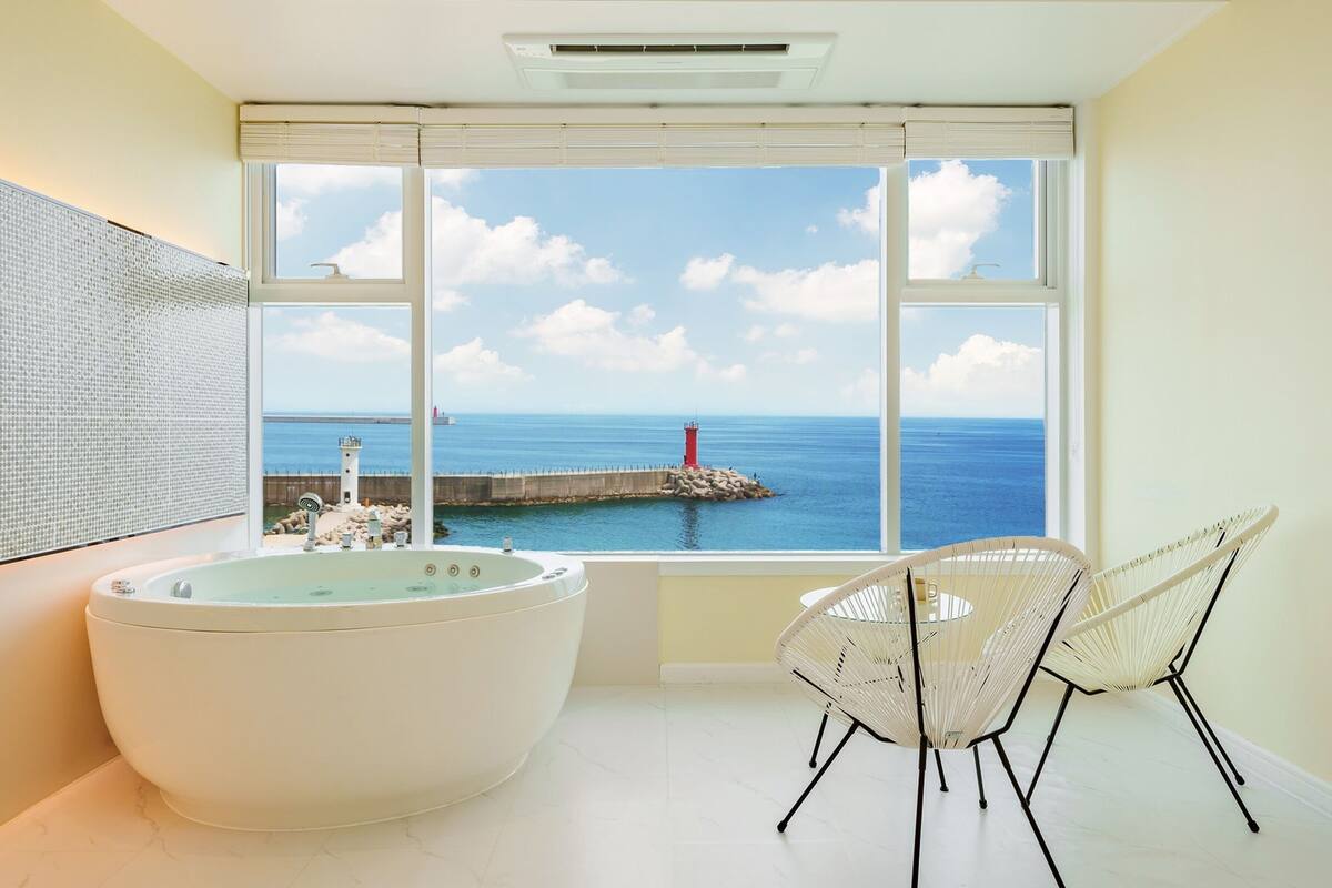 Un baño con una bañera grande, una silla y vista al mar a través de un ventanal amplio.