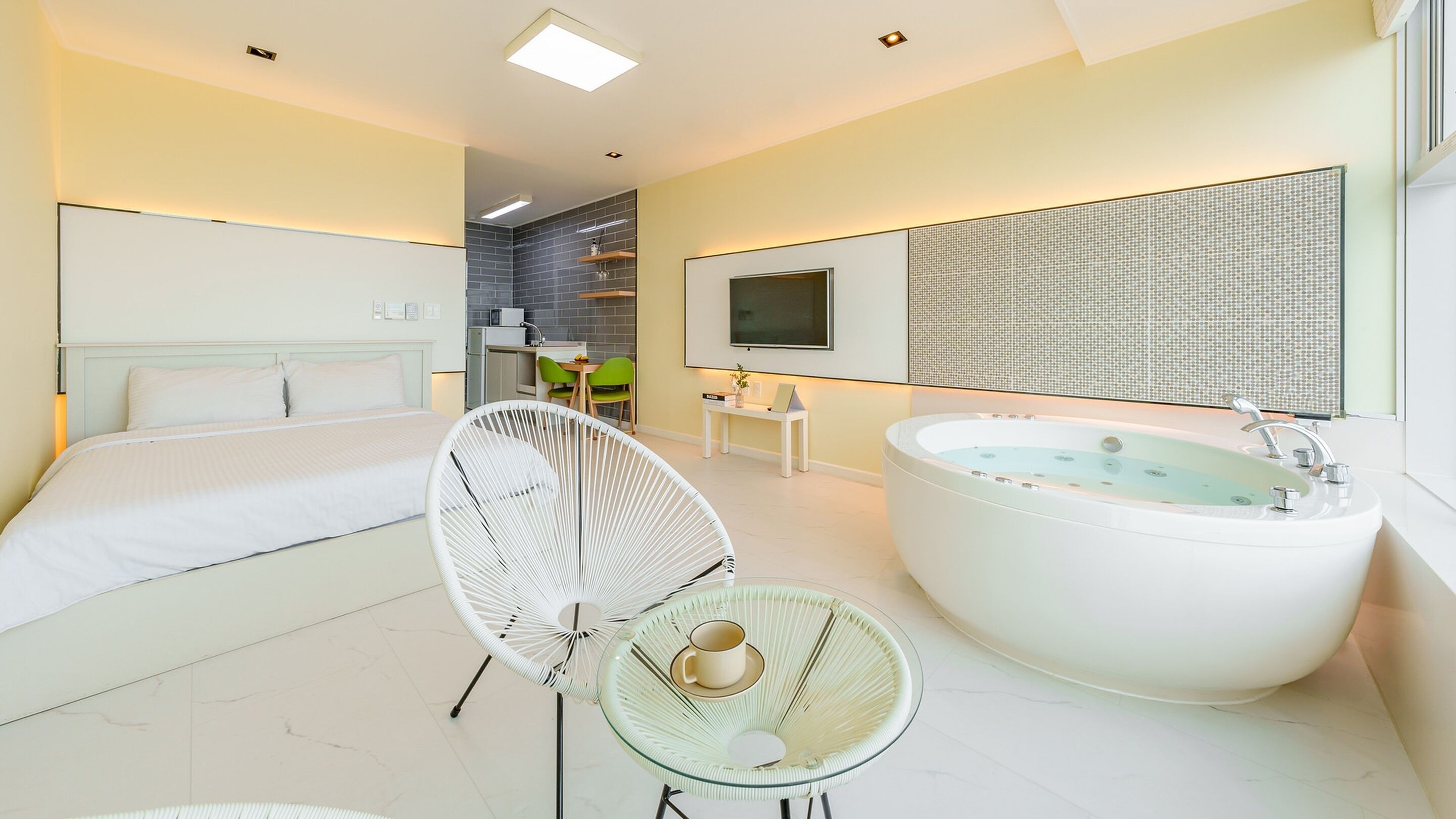 A modern hotel room with a large bed, a glass chair, a bathtub, and a small kitchenette.