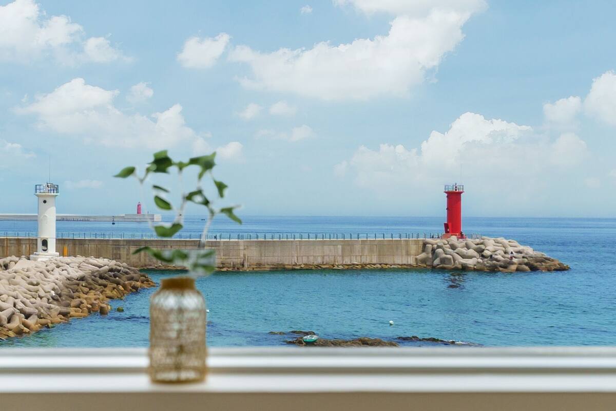 Vista costera con dos faros, uno blanco y otro rojo, y un rompeolas rocoso.