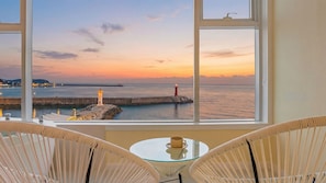 Ein Zimmer mit Blick auf einen Sonnenuntergang über dem Meer, mit einem Leuchtturm, einer Pier und einem Glastisch mit Stühlen.