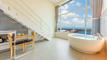 Un baño moderno con una bañera grande, una zona de comedor con paredes de vidrio y una escalera que lleva a un balcón con vistas al mar.