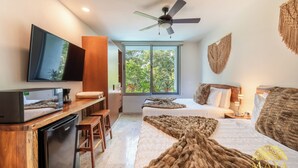 A modern hotel room with a flat-screen TV, a ceiling fan, a large window with a view of trees, a bed with a brown blanket, and a wooden desk with a coffee maker.