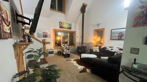 Ein gemütliches Wohnzimmer mit Kamin, einer Couch, einem Sessel und einem Couchtisch. Es gibt ein Fenster mit Vorhängen, eine Pflanze und einen an der Wand befestigten Fernseher.