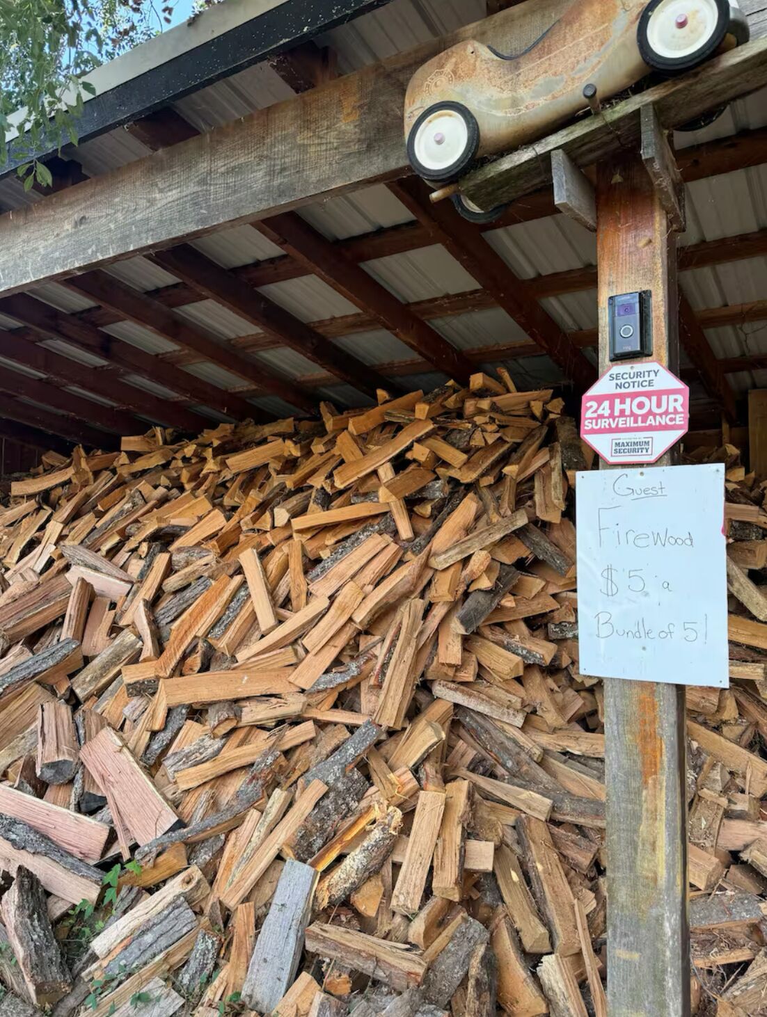 Une pile de bois de chauffage, avec un panneau indiquant que la zone est sous surveillance 24 heures sur 24 et que le prix est de 5 dollars par paquet de 5 bûches.