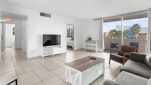 Un salon moderne avec un téléviseur à écran plat, une table basse et un canapé. Il y a un balcon avec vue sur les palmiers et les immeubles.