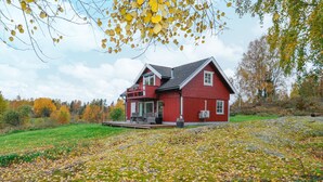 Une maison rouge avec un balcon, entourée d’un champ verdoyant et d’arbres aux feuilles d’automne.