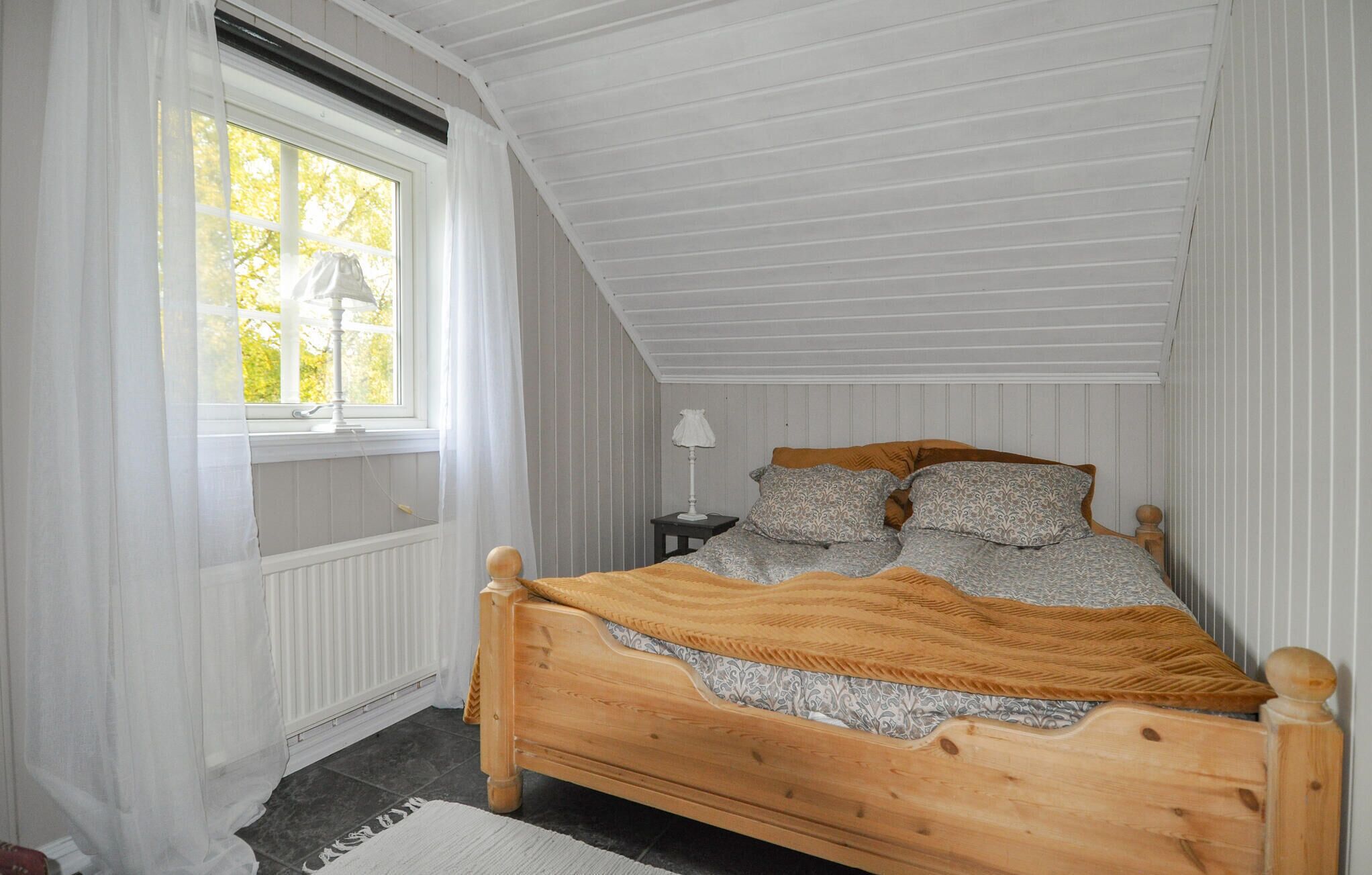A bedroom with a wooden bed, a window with curtains, a radiator, and a lamp on a bedside table.