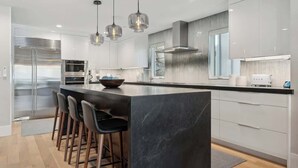 Une cuisine moderne dotée d’un îlot central, de tabourets de bar, d’appareils électroménagers en acier inoxydable, de luminaires suspendus et d’une grande fenêtre.