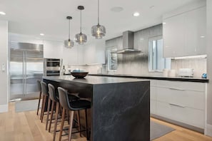 Une cuisine moderne dotée d’un îlot central, de tabourets de bar, d’appareils électroménagers en acier inoxydable, de luminaires suspendus et d’une grande fenêtre.