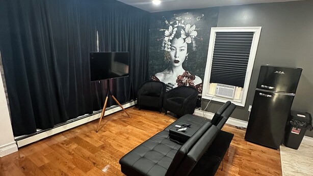 A modern living room with a black sectional sofa, a flat-screen TV, a large mural, and a black refrigerator.