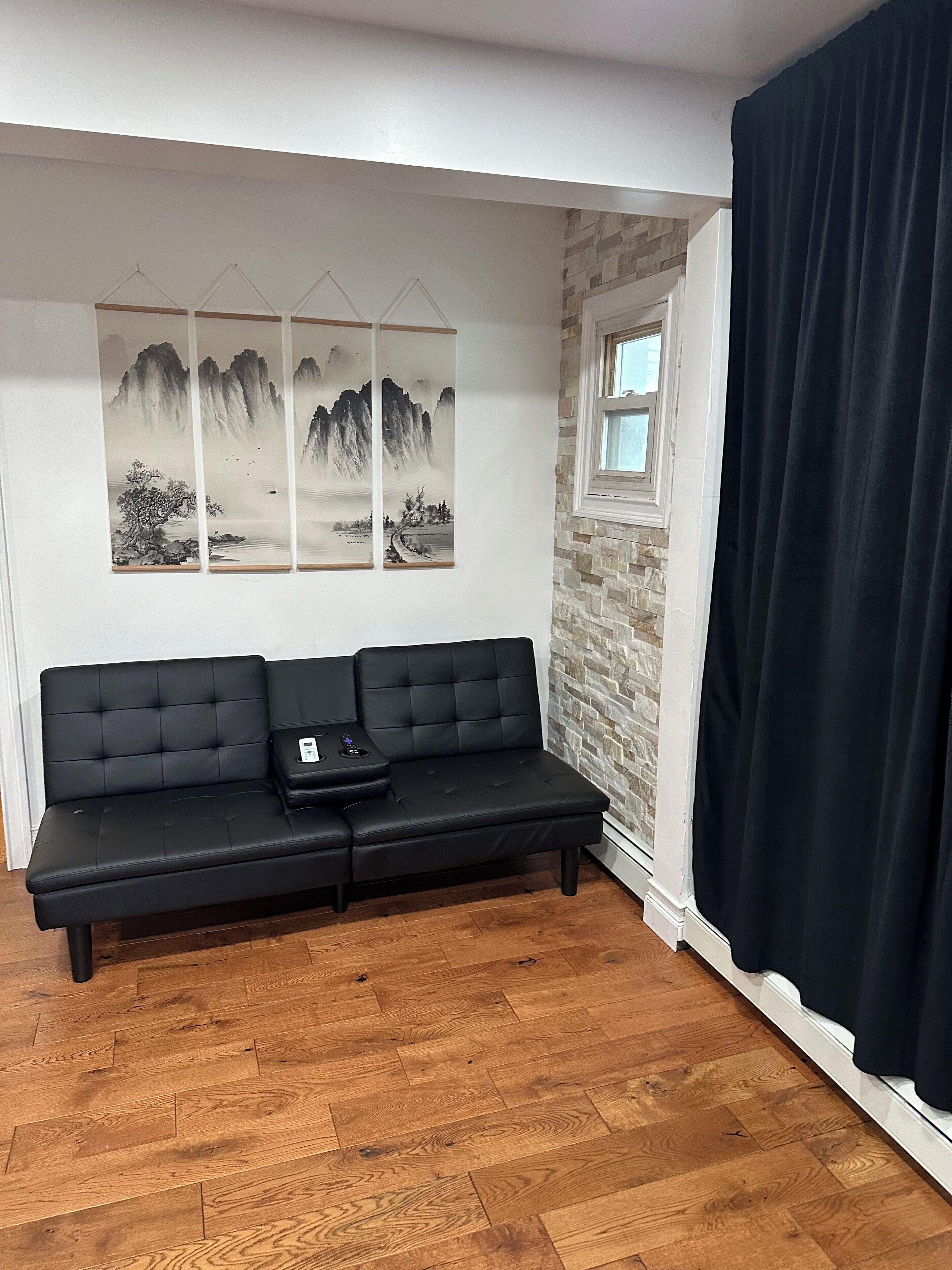 A black leather sofa with a remote control, a wooden floor, a stone wall, and a window with a curtain.