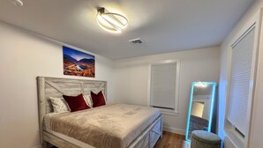 A bedroom with a wooden bed, a nightstand, a mirror with LED lights, a window with blinds, and a painting on the wall. - Family Vacation Modern Norwood  (Norwood)