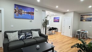 A modern living room with a dark grey sofa, a black coffee table, and a grey patterned rug. There are two paintings on the walls, a dining area with a small table and chairs, and a wooden floor. - Family Vacation Modern Norwood  (Norwood)