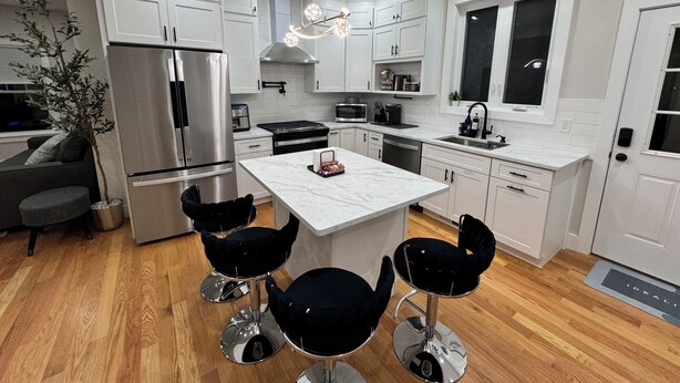 A modern kitchen with white cabinetry, a stainless steel refrigerator, a central island with bar stools, and a chandelier. - Family Vacation Modern Norwood  (Norwood)