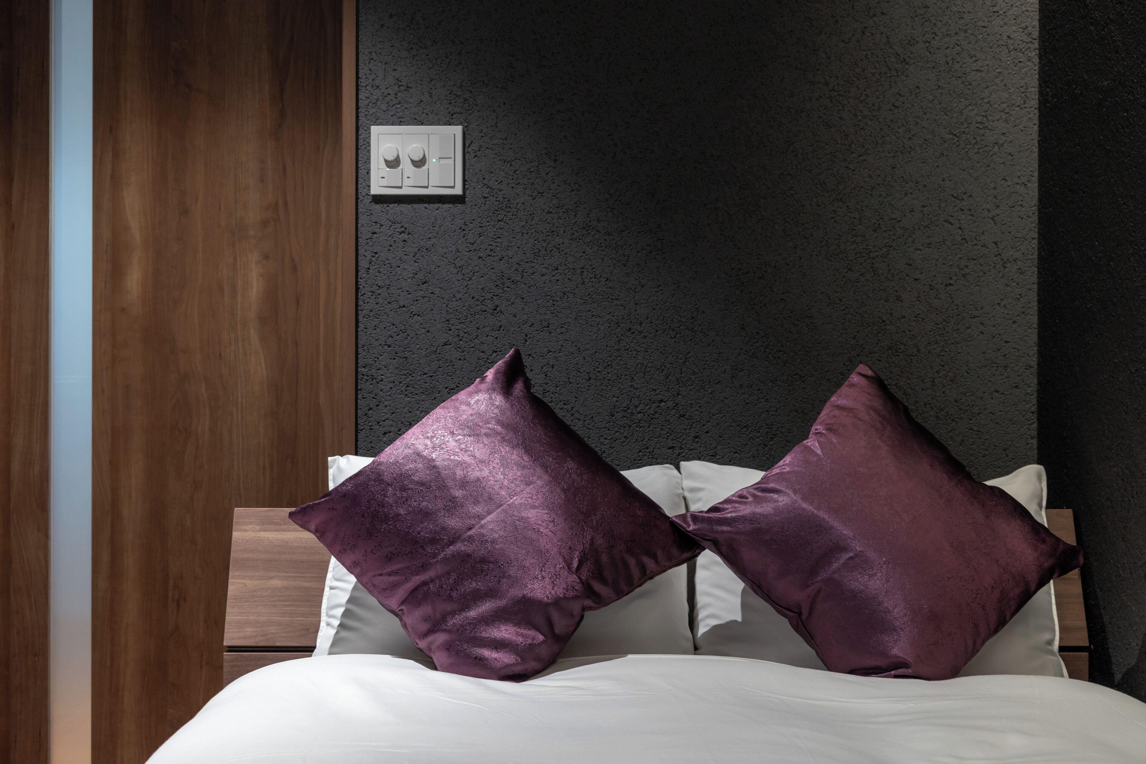 A bed with white linens and two purple pillows, a wooden headboard, and a textured dark wall.