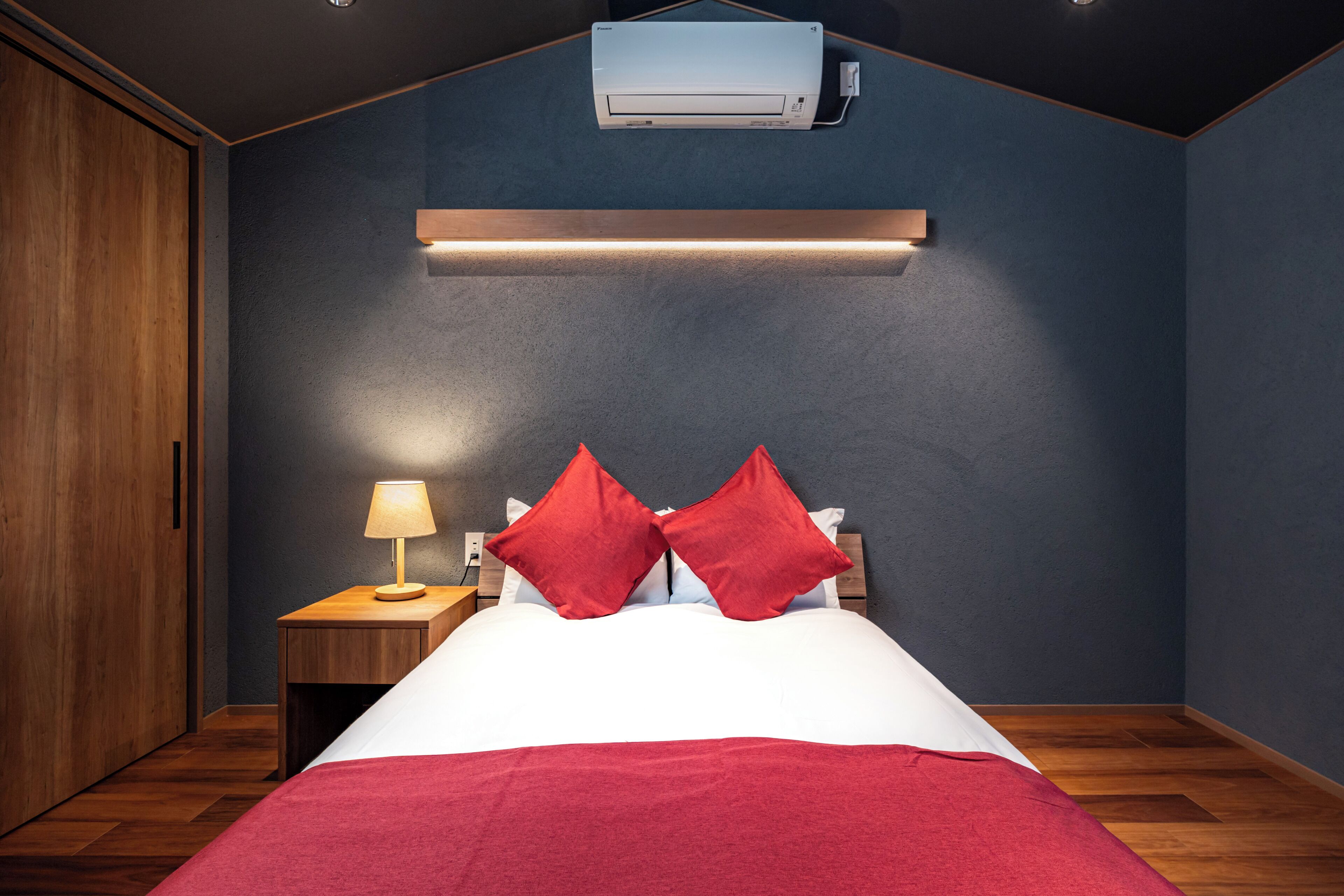 A bedroom with a bed, red pillows, a nightstand with a lamp, a wooden wardrobe, and an air conditioning unit on the wall.