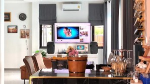 A modern living room with a large flat-screen TV, a dining table with chairs, a coffee table, a fireplace, and a bookshelf. - Cher Cher Pool villa (Ban Chang)