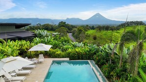 A pool area with lounge chairs and an umbrella, surrounded by lush greenery and a view of a mountain. - Serene Hills Arenal (Monterrey)
