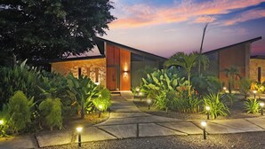 A modern building with a stone facade and a wooden door. There is a pathway leading to the entrance, surrounded by lush greenery and illuminated by ground lights. - Serene Hills Arenal (Monterrey)