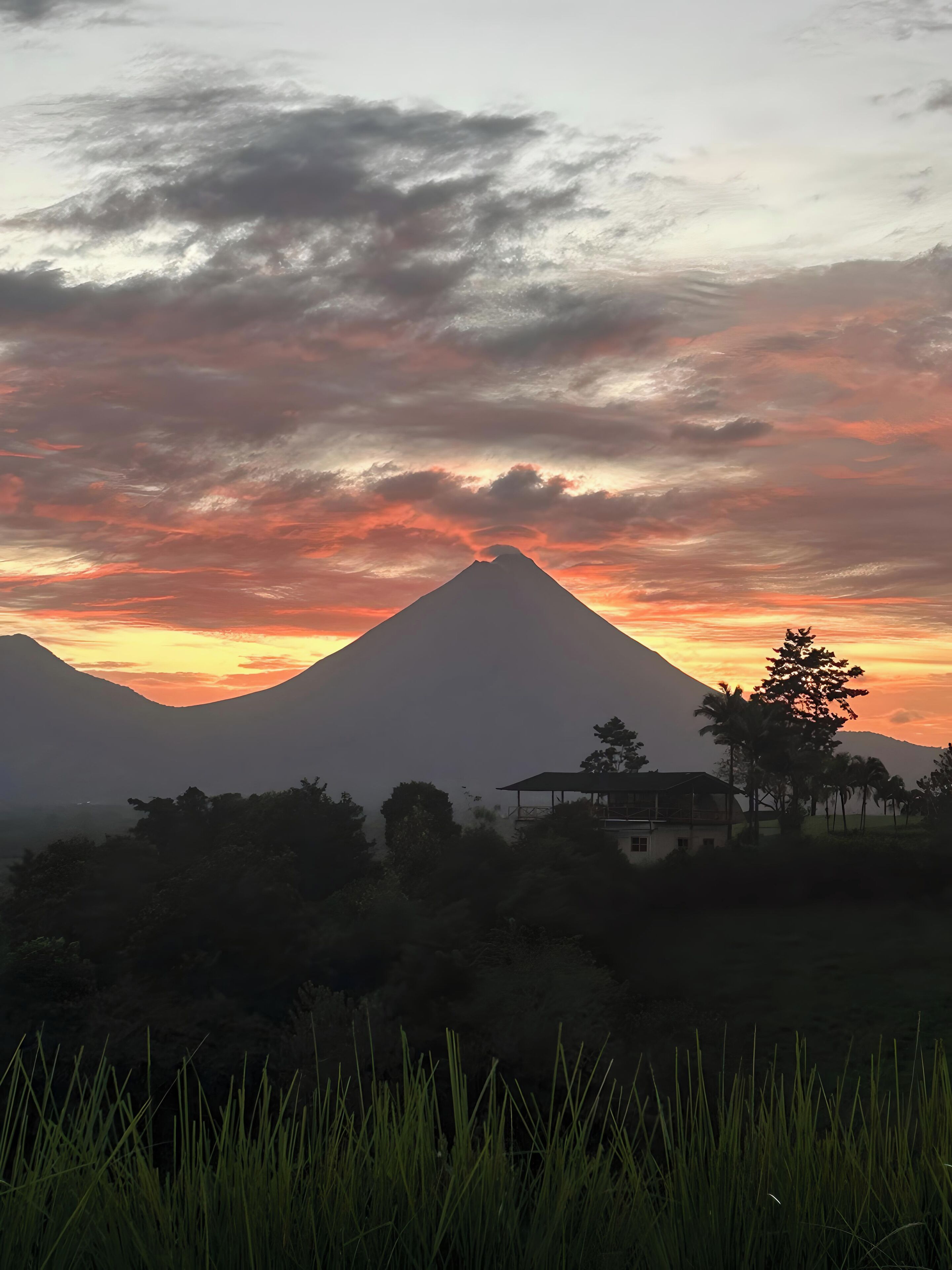 A mountain with a peak silhouetted against a sunset sky, surrounded by greenery and a small building.