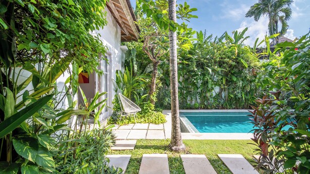 A poolside area with a clear blue pool, surrounded by lush greenery and a white building with a tiled roof.