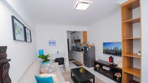Uma sala moderna com sofá, mesa de centro e televisão. Cozinha visível ao fundo, com armários e geladeira.