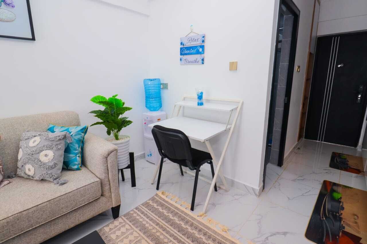 Un espace de vie compact comprenant un canapé, une petite table, une chaise, une plante en pot, un distributeur d’eau et un panneau affichant des mots d’encouragement.