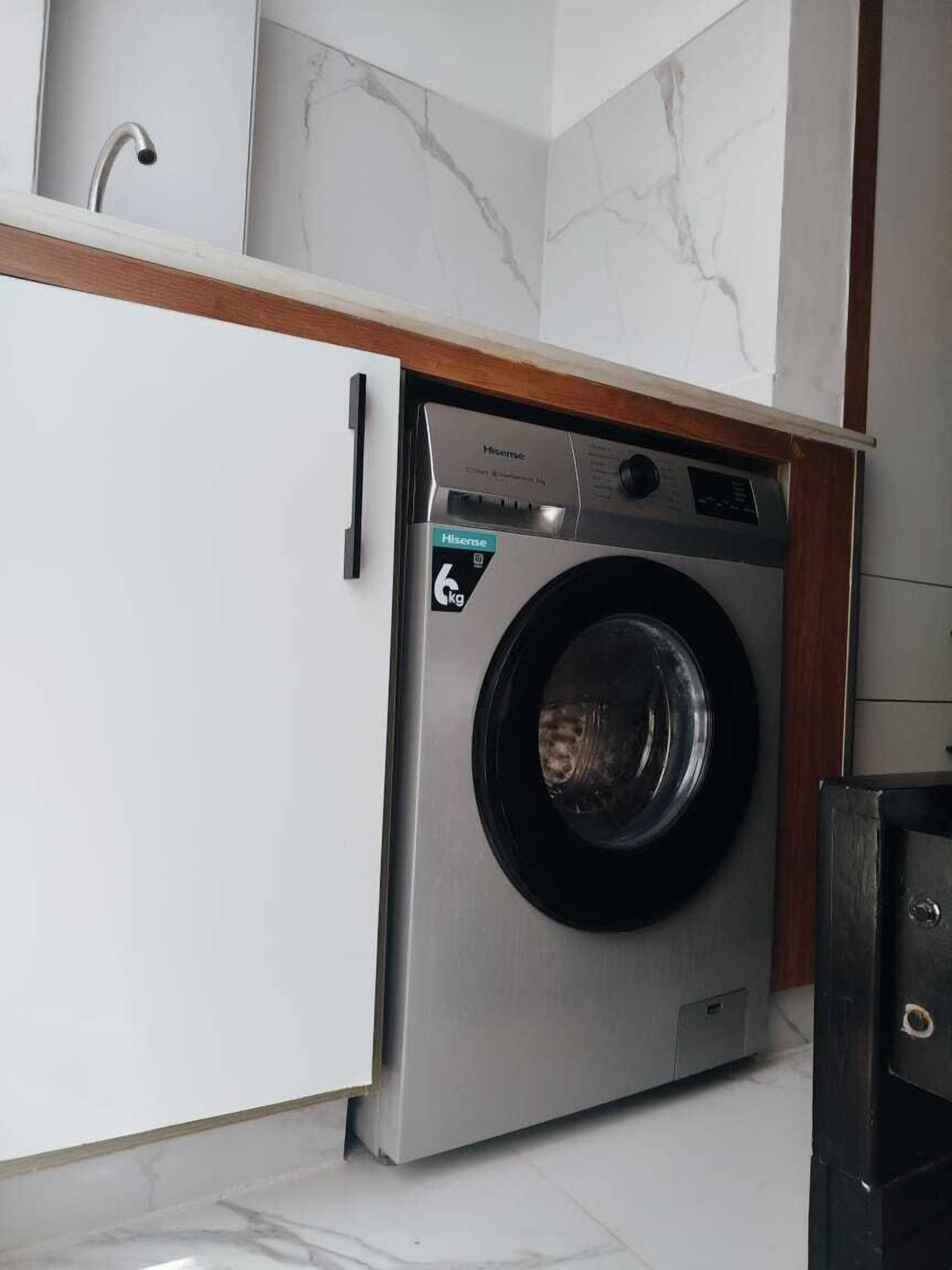 A compact, stainless steel front-loading washing machine with a control panel, placed in a modern kitchen with white cabinetry and a marble backsplash.