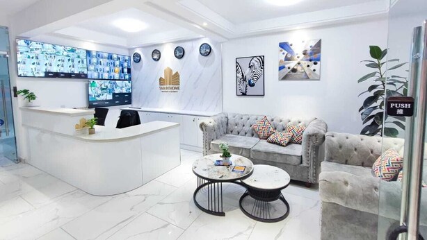 Uma área de recepção moderna com um balcão branco, um sofá cinza com almofadas estampadas e duas mesas redondas.