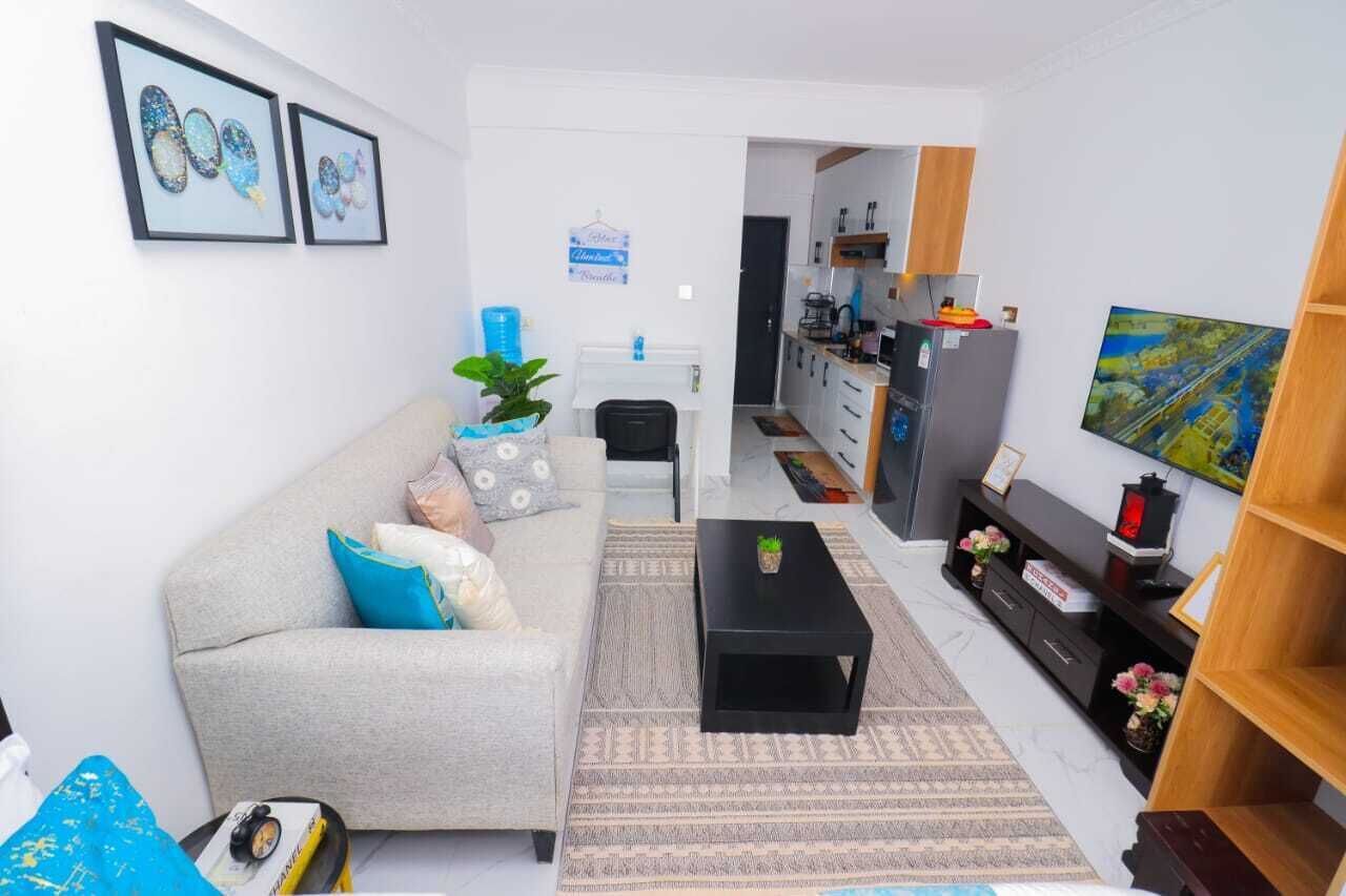 A living room with a sofa, coffee table, and a TV on the wall. There is a kitchen in the background with a fridge, microwave, and a painting.