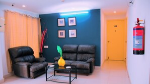 A living room with two leather sofas, a coffee table, and a vase with a plant. There are framed pictures on the wall and a fire extinguisher mounted on the wall. - Naksha Tree Hotels Honey Crest (Chennai)