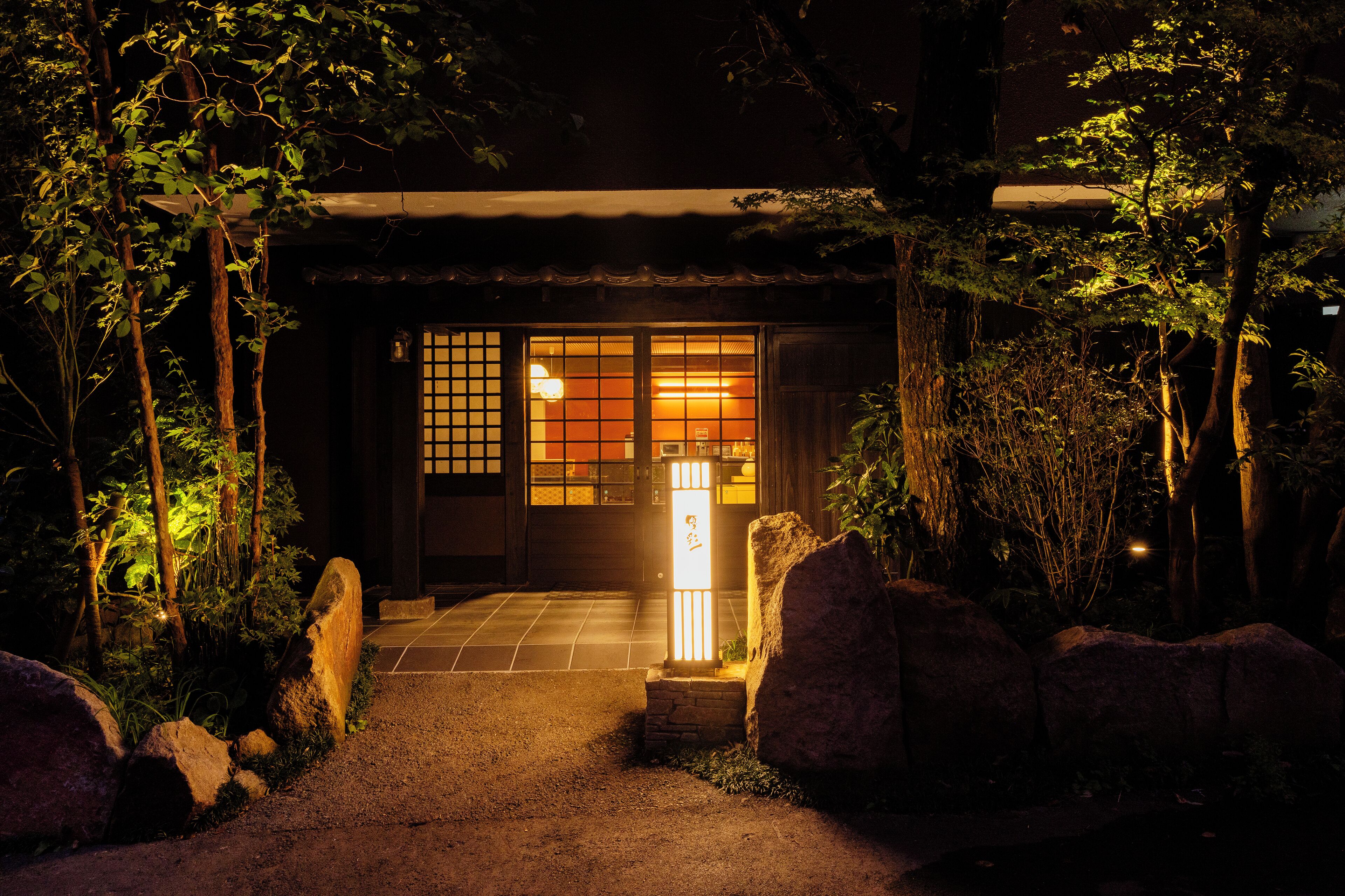 A traditional Japanese building entrance at night with a lit lantern, surrounded by rocks and foliage.