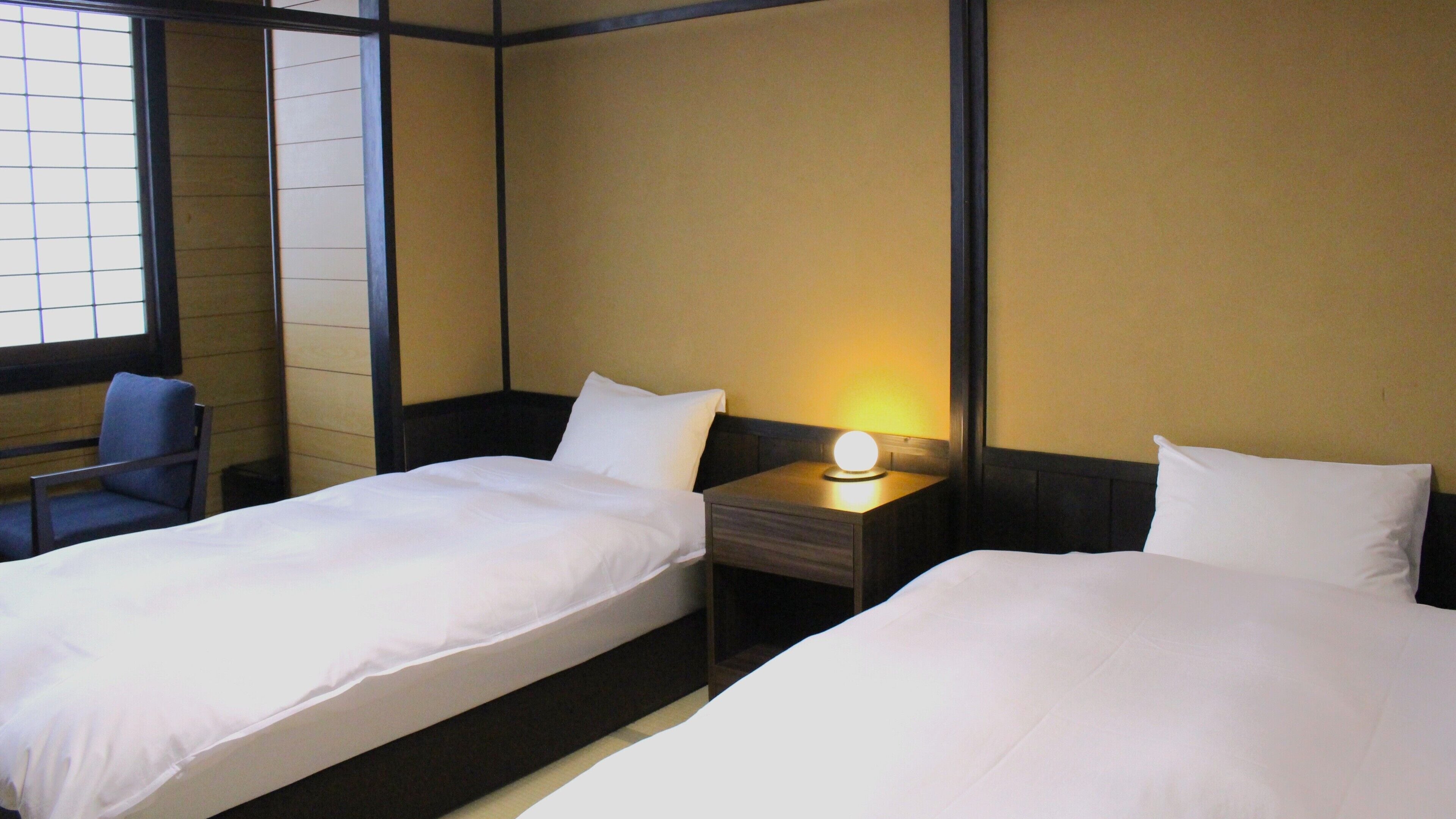 A hotel room with two beds, a nightstand with a lamp, a chair, and a window with blinds.
