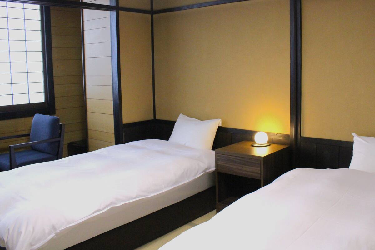 Una camera d'albergo con due letti, un comodino con lampada, una sedia e una finestra con tende.