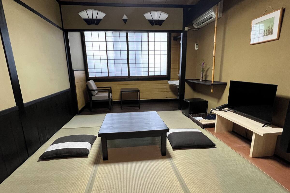 Una stanza tradizionale con tatami, un tavolino basso, cuscini, una televisione e una finestra con paraventi shoji.