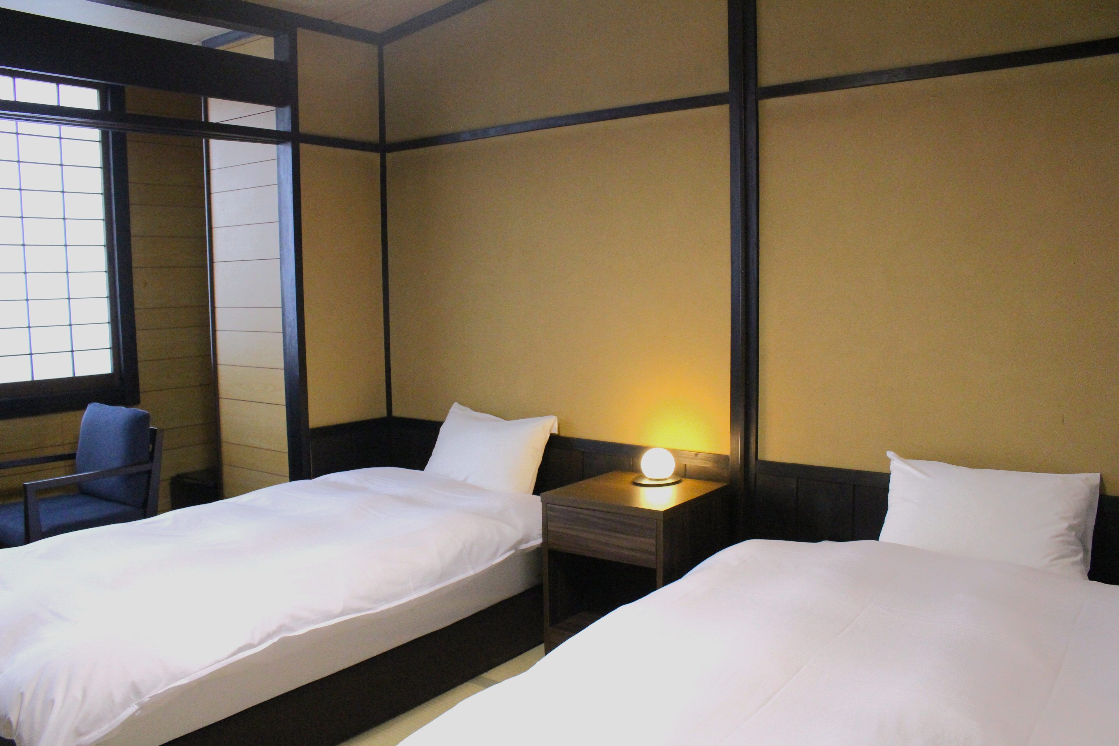 Habitación de hotel con dos camas, una silla y una mesita de noche con lámpara.