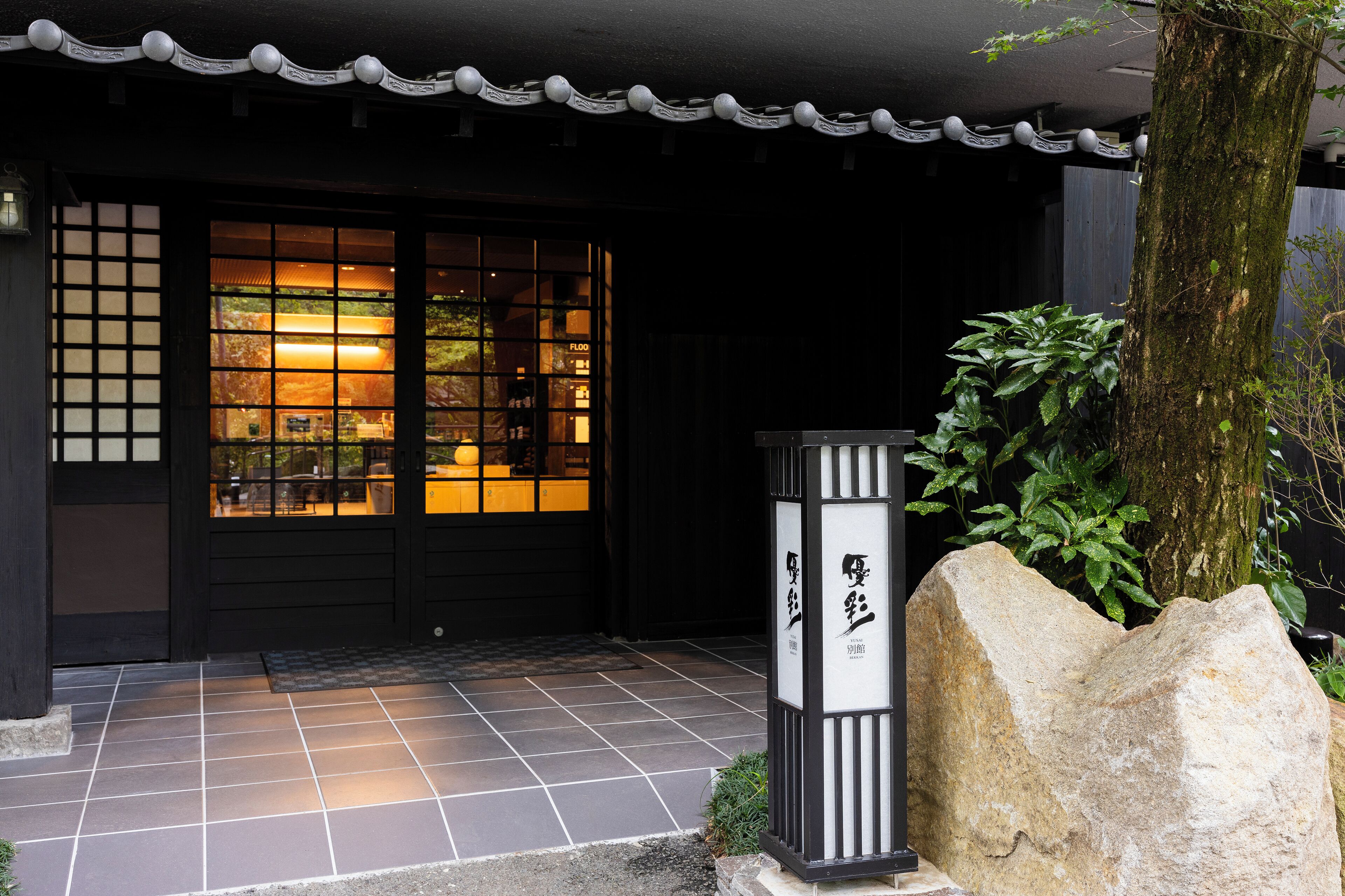 A traditional Japanese building entrance with a tiled floor, a black door with glass panels, and a stone lantern.