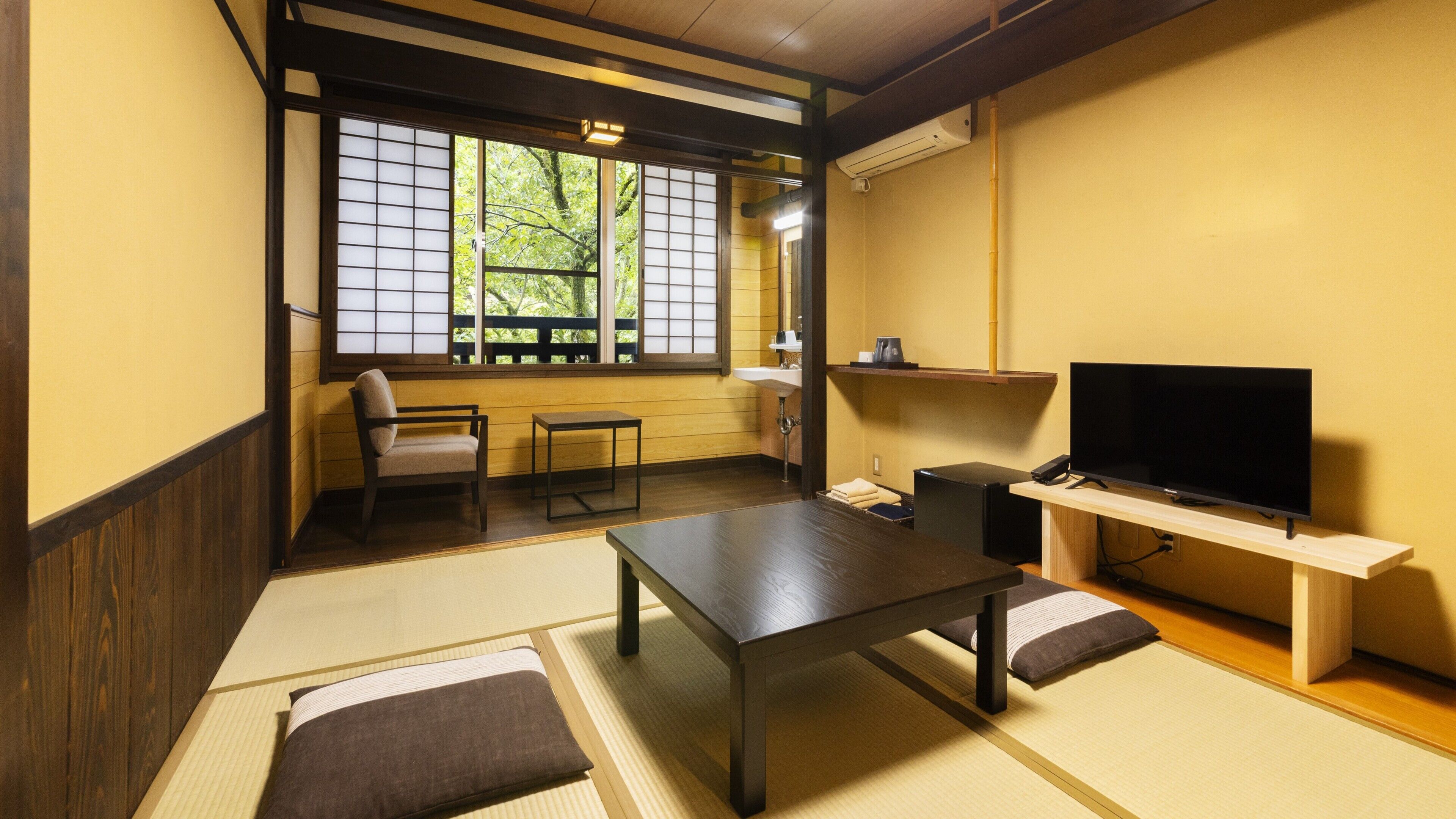 A traditional Japanese-style room with tatami flooring, a low table, a sofa, a television, and a window with a view of trees.