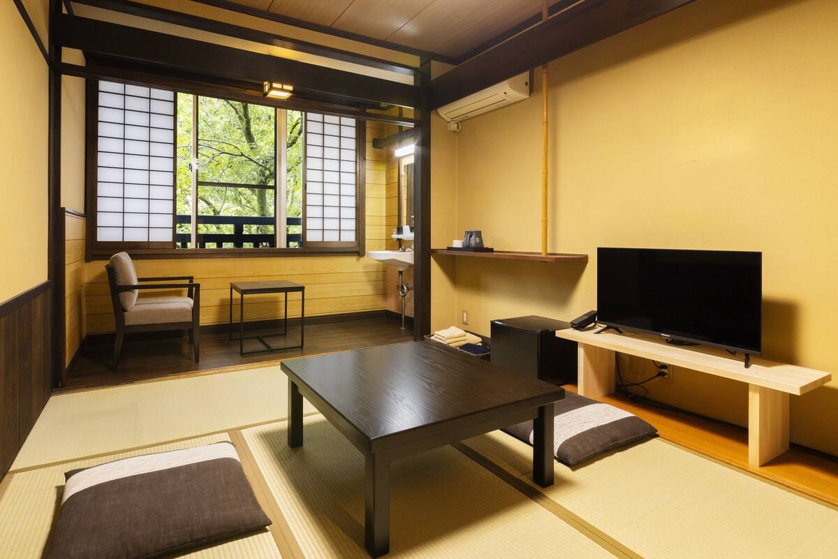 Una camera tradizionale in stile giapponese, con pavimento in tatami, un tavolino, una poltrona e una televisione, e una finestra vista alberi.