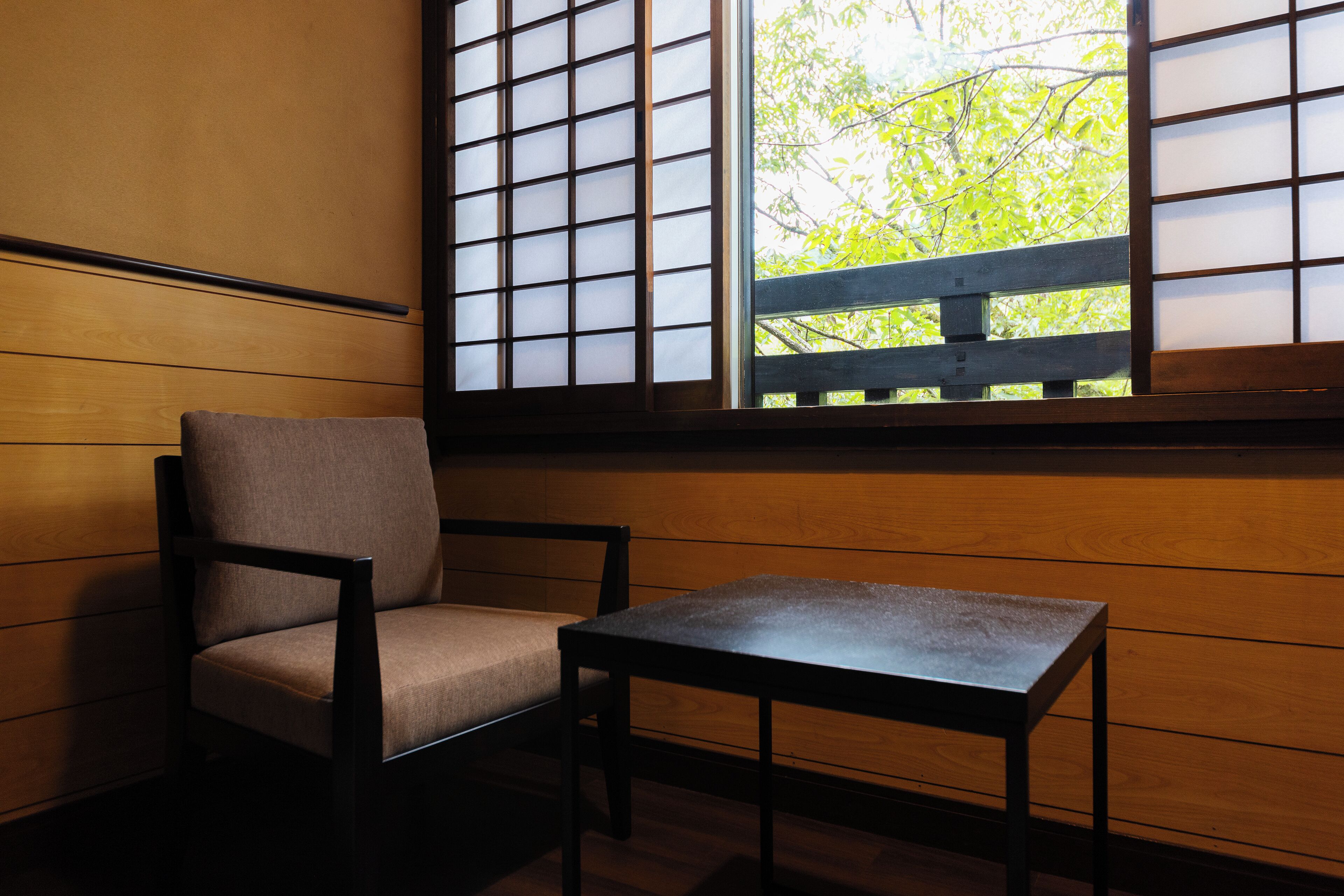 Una habitación con pared de madera, una ventana con vista a vegetación, una silla y una pequeña mesa.