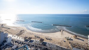 Zona costera con playa, una carretera con coches aparcados y un edificio de techo plano.
